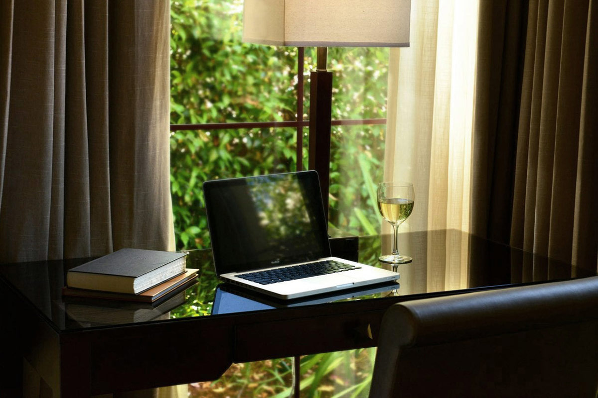 A laptop, a glass of white wine, two books placed on a desk in a guest accommodation at Vintners Resort. The desk is placed by the window overlooking the garden.
