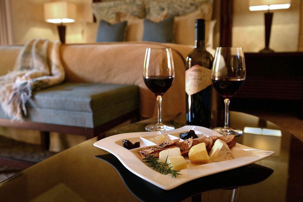 In-room dining at Vintners Resort: a plate of cheese and crackers with two glasses of red wine and a bottle of wine, against the backdrop of a guestroom.