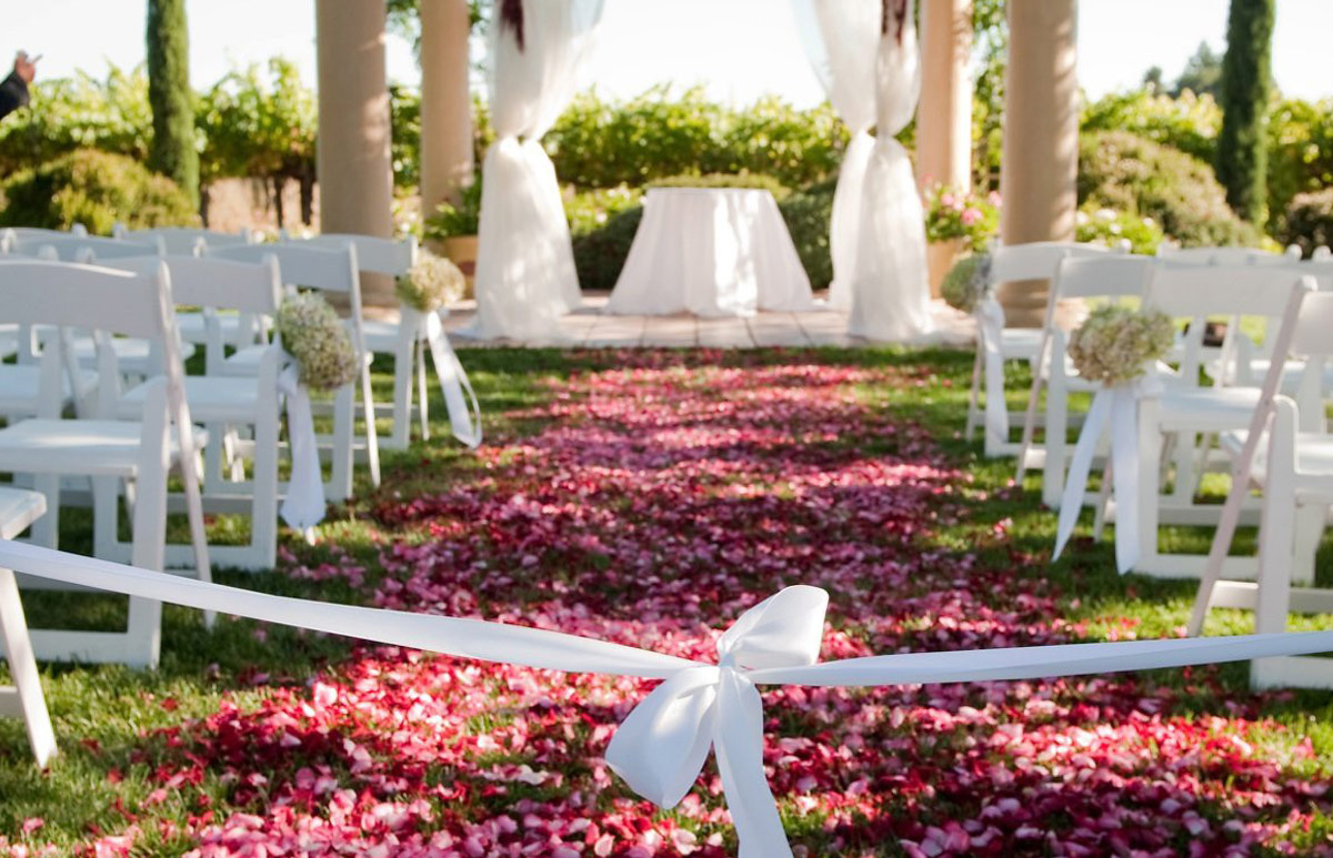 An outdoor wedding ceremony set up at Vintners Resort's Fontana Pavilion, with a white ribbon at the entrance, white chairs on both sides, and the aisle is covered with red flower pedals.