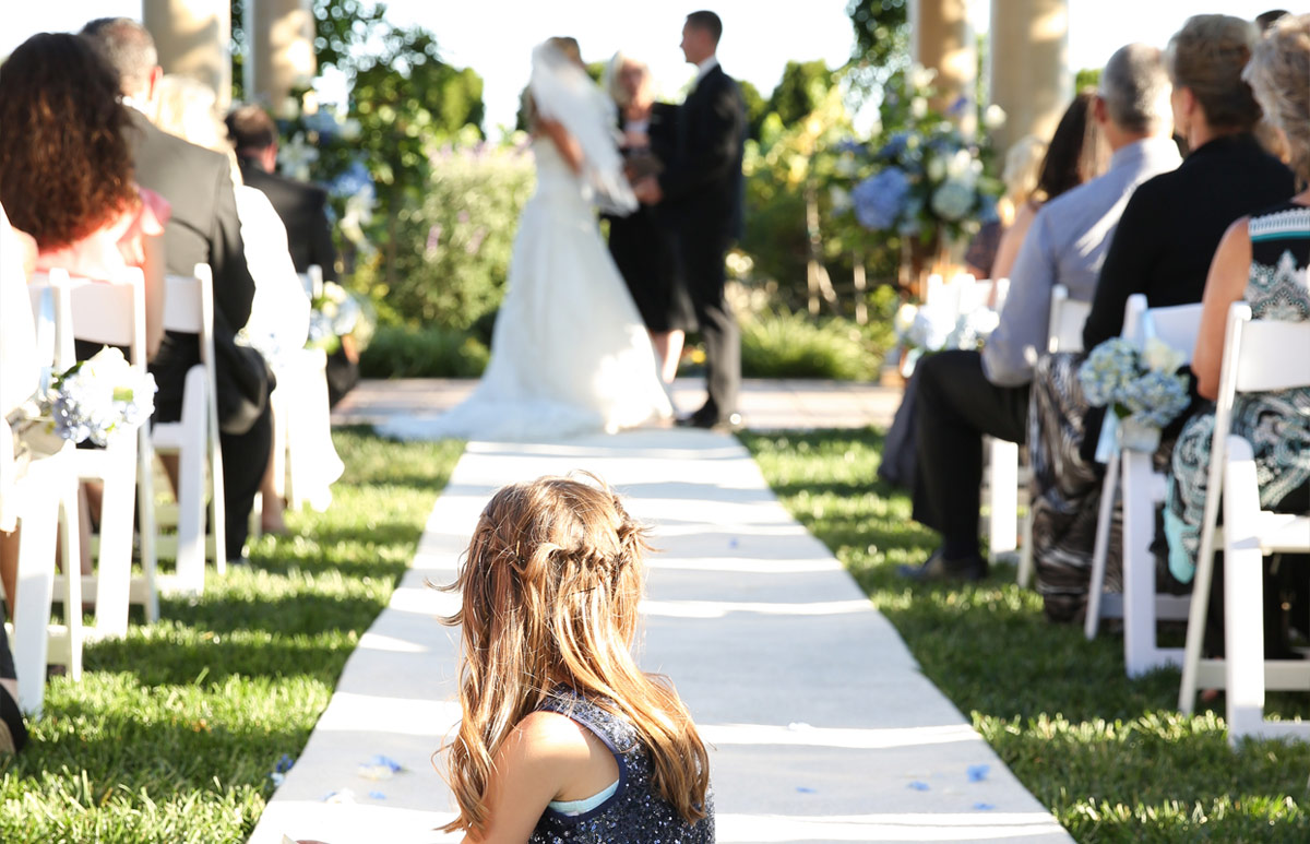 An outdoor wedding ceremony set up at Vintners Resort. A little girl is sitting on the white carpeted aisle, guessts sitting on both sides of the aisle, and the bride and groom under the Fontana Pavilion.