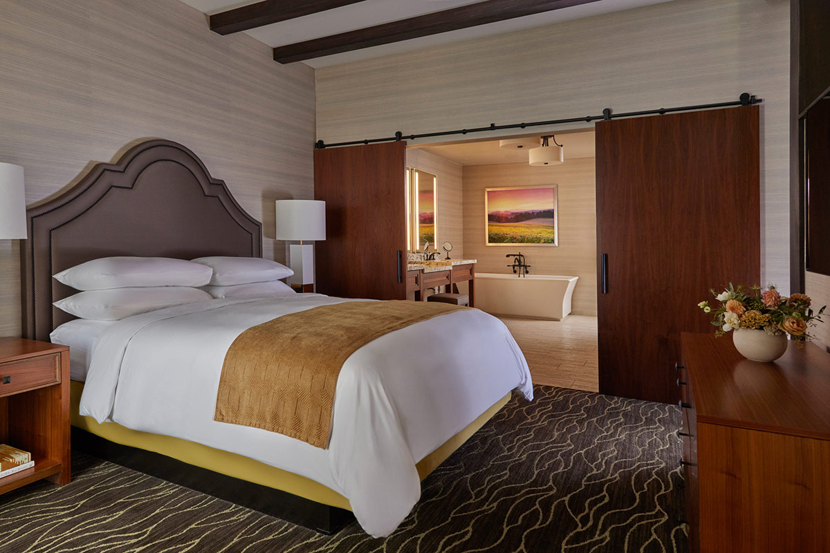 The bedroom of the Vineyard View Suite featues a comfortable California king feather bed with plush down bedding, looking into the spacious bathroom with a Roman soaking tub and double vanity.